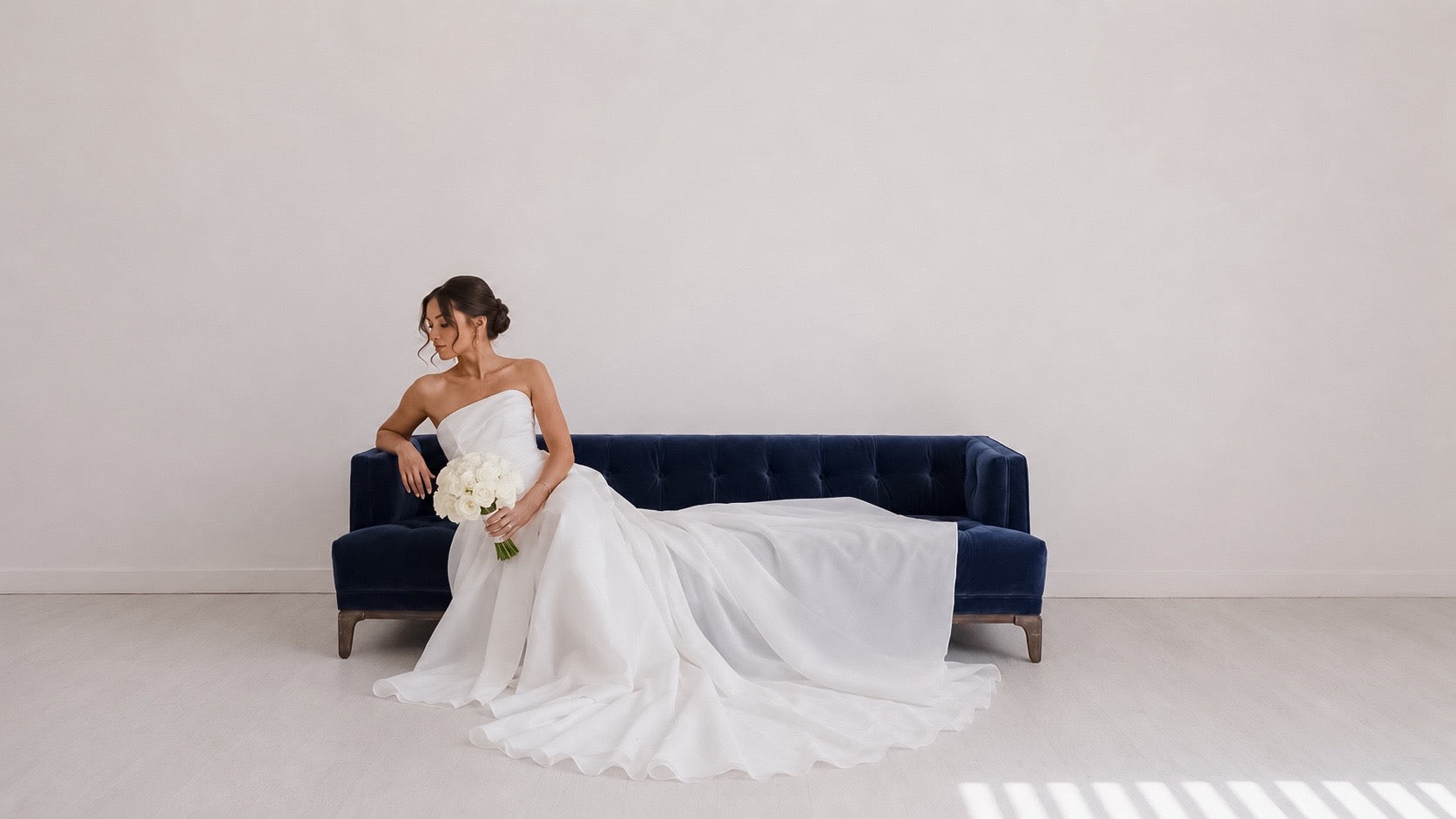 Bride in a white wedding gown seated on a navy velvet sofa holding a white rose bridal bouquet